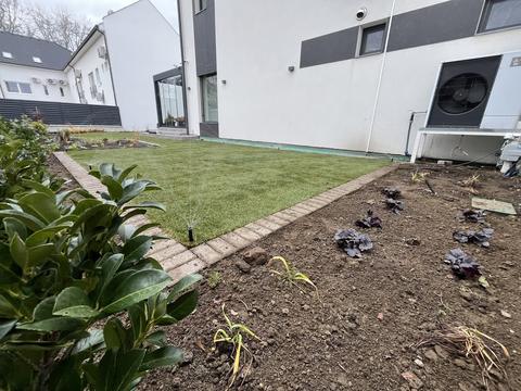 Frissen fektetett gyepszőnyeg klinkertégla szegéllyel egy modern ház mellett, a gyep sarkában talajszinti szórófejjel, előtérben frissen ültetett évelőkkel a csupasz földben