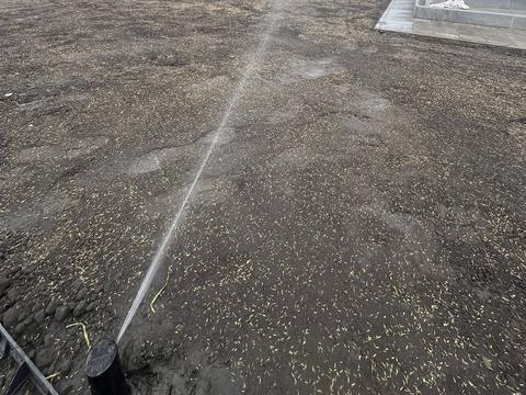 Rotoros szórófej a kép alsó szélén, egyetlen összefüggő vízsugárral szórja a frissen elmunkált, vetésre előkészített földfelületet, háttérben ház, kerítés és felhős ég